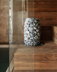 Wooden sauna with a stack of stones inside, surrounded by wooden benches and walls and a glass wall next to a pool