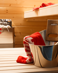Metal bucket with a red and white towel and wooden bench in a wooden sauna. Harvia The Wall Sauna Heater Stainless Steel mounted on sauna wall in background