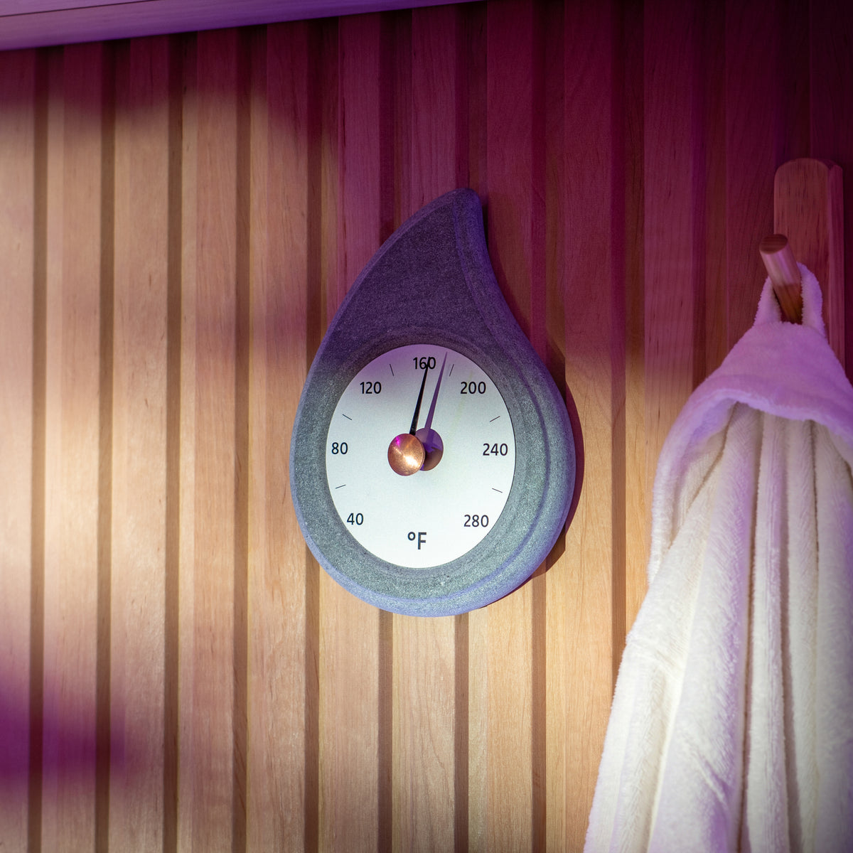 stone teardrop shaped sauna thermometer with a brass knob hanging on a wooden wall with a robe on a hook