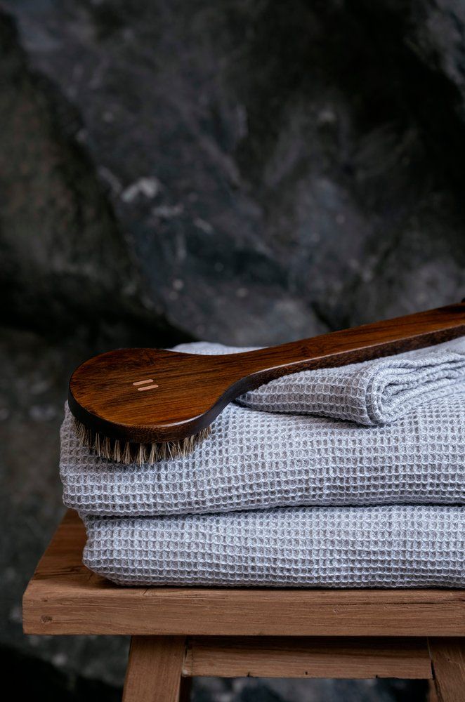A folded light gray waffle-weave towel or blanket sits on a wooden stool, with a dark brown wooden scrubber resting across the top, set against a dark, textured stone background.