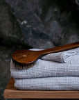 A folded light gray waffle-weave towel or blanket sits on a wooden stool, with a dark brown wooden scrubber resting across the top, set against a dark, textured stone background.
