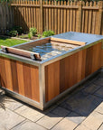 A rectangular outdoor hot tub or plunge pool with a wood-look exterior and stainless steel trim, shown on a stone patio in a backyard with a wooden fence and grass.