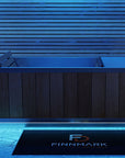 A modern rectangular cold plunge with a dark wood-paneled front and light blue interior, shown in a blue-lit bathroom setting with a black FINMARK DESIGNS floor mat in front.