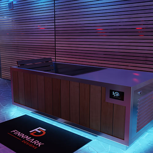 A modern cold plunge tub with a wood-paneled cabinet and stainless steel top, shown in a pool area with blue and purple ambient lighting. A black Finnmark Designs branded mat is placed in front.