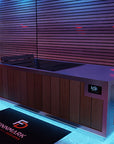 A modern cold plunge tub with a wood-paneled cabinet and stainless steel top, shown in a pool area with blue and purple ambient lighting. A black Finnmark Designs branded mat is placed in front.