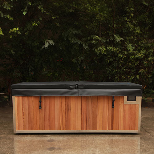 A rectangular cold plunge tub with a warm brown wood-look front, light beige sides, and a dark gray cover, photographed outdoors against a background of green foliage and a concrete ground.