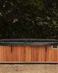 A rectangular cold plunge tub with a warm brown wood-look front, light beige sides, and a dark gray cover, photographed outdoors against a background of green foliage and a concrete ground.
