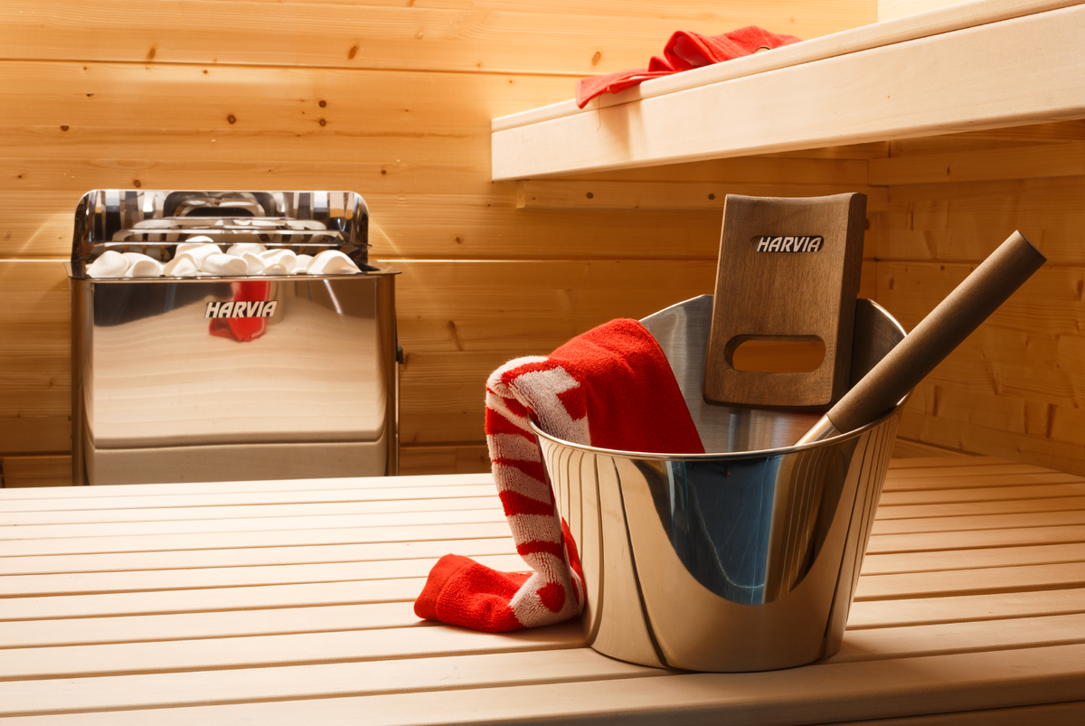 Metal bucket with a red and white towel and wooden bench in a wooden  sauna. Harvia The Wall Sauna Heater Stainless Steel mounted on sauna wall in background