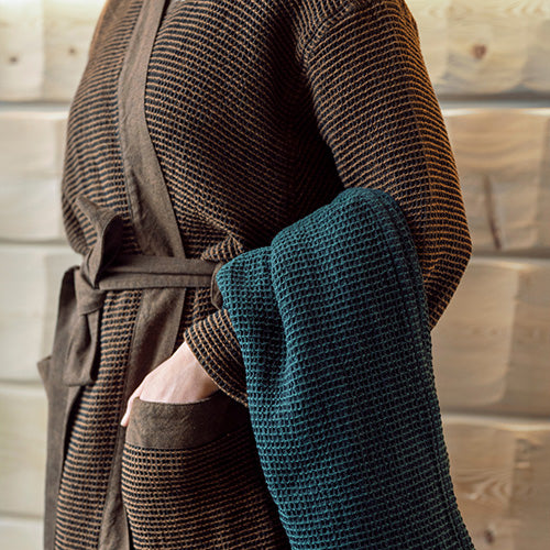 Person wearing a brown textured sauna robe holding a blue towel against a neutral background