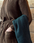 Person wearing a brown textured sauna robe holding a blue towel against a neutral background