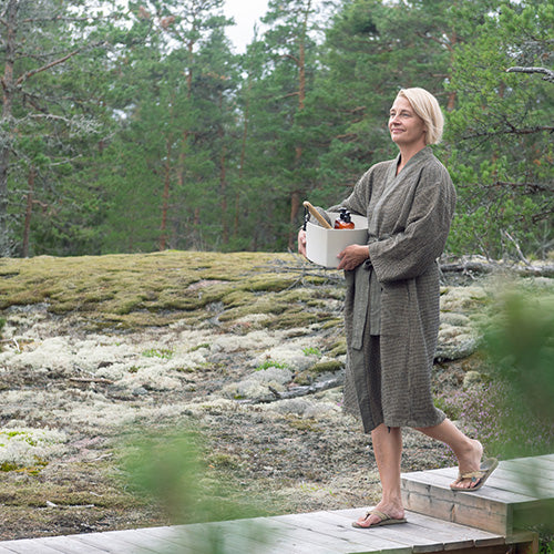 A woman in a textured brown sauna bathrobe walks along a wooden boardwalk in a mossy forest clearing, holding a white cooler with bottles.