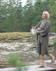 A woman in a textured brown sauna bathrobe walks along a wooden boardwalk in a mossy forest clearing, holding a white cooler with bottles.