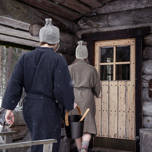 Two people wearing woven sauna robes and knit hats walk toward a rustic log cabin door, one carrying a black bucket with a wooden spoon. Snow is lightly visible in the air, and the cabin wood and door frame the background.