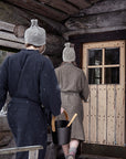 Two people wearing woven sauna robes and knit hats walk toward a rustic log cabin door, one carrying a black bucket with a wooden spoon. Snow is lightly visible in the air, and the cabin wood and door frame the background.