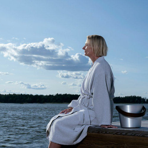 A woman in profile sits on a wooden dock by the water wearing a light grey textured sauna robe, with a silver metal bucket beside her under a blue sky with clouds.