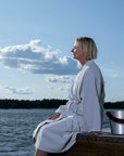 A woman in profile sits on a wooden dock by the water wearing a light grey textured sauna robe, with a silver metal bucket beside her under a blue sky with clouds.