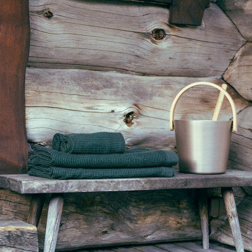 folded dark green waffle knit towels on a rustic wooden bench next to an aluminum bucket with light wood handle and ladle against a wooden cabin background