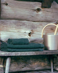 folded dark green waffle knit towels on a rustic wooden bench next to an aluminum bucket with light wood handle and ladle against a wooden cabin background