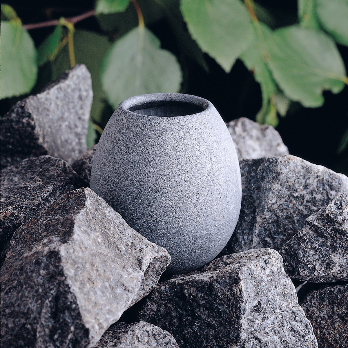 light grey sauna stone aroma cup sitting amongst dark grey heater stones and leaves in the background
