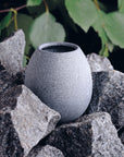 light grey sauna stone aroma cup sitting amongst dark grey heater stones and leaves in the background