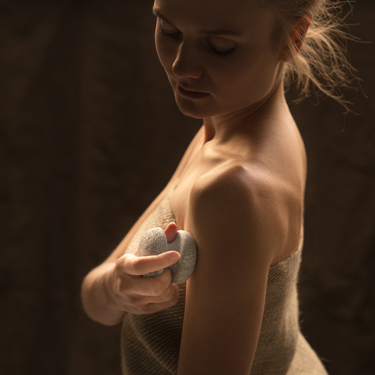woman hold a gray sauna stone with two round knob shaped ends and touching her arm with it