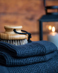 folded dark grey textured towels with a bristle brush on top with a blurred background and lit lantern and candle