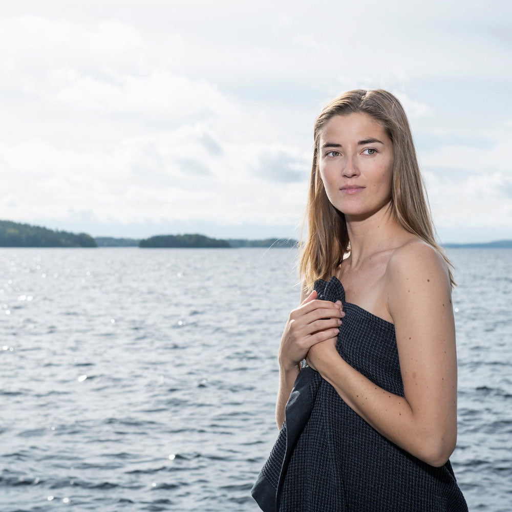 women standing with dark grey textured towel wrapped around her with a lake behind her and trees in the distance